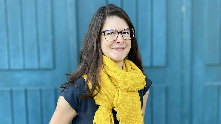 Portraitfoto von Karin Hediger vor einem blauen Hintergrund. Sie trägt einen gelben Schal, hat schulterlange braune Harre und eine Brille.